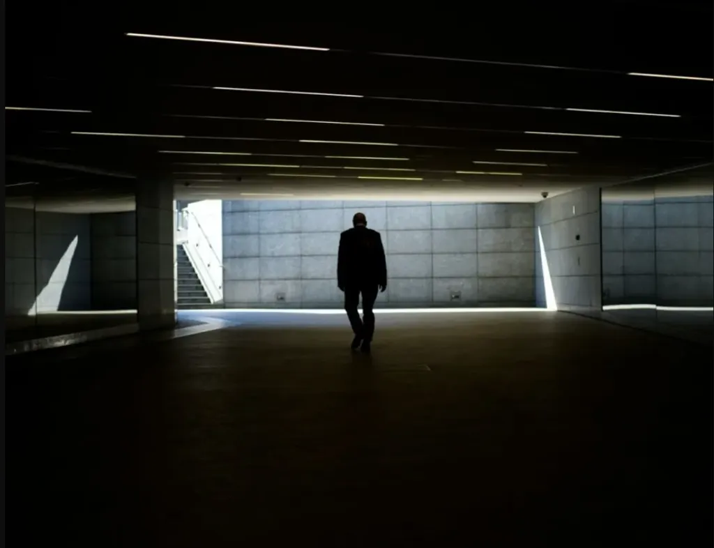A silhouetted figure in a suit walks alone through a dark underground corridor toward a bright light at the far end.