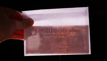 A hand holds up Hungarian currency in a white envelope up to the light. The currency is visible from inside the envelope.
